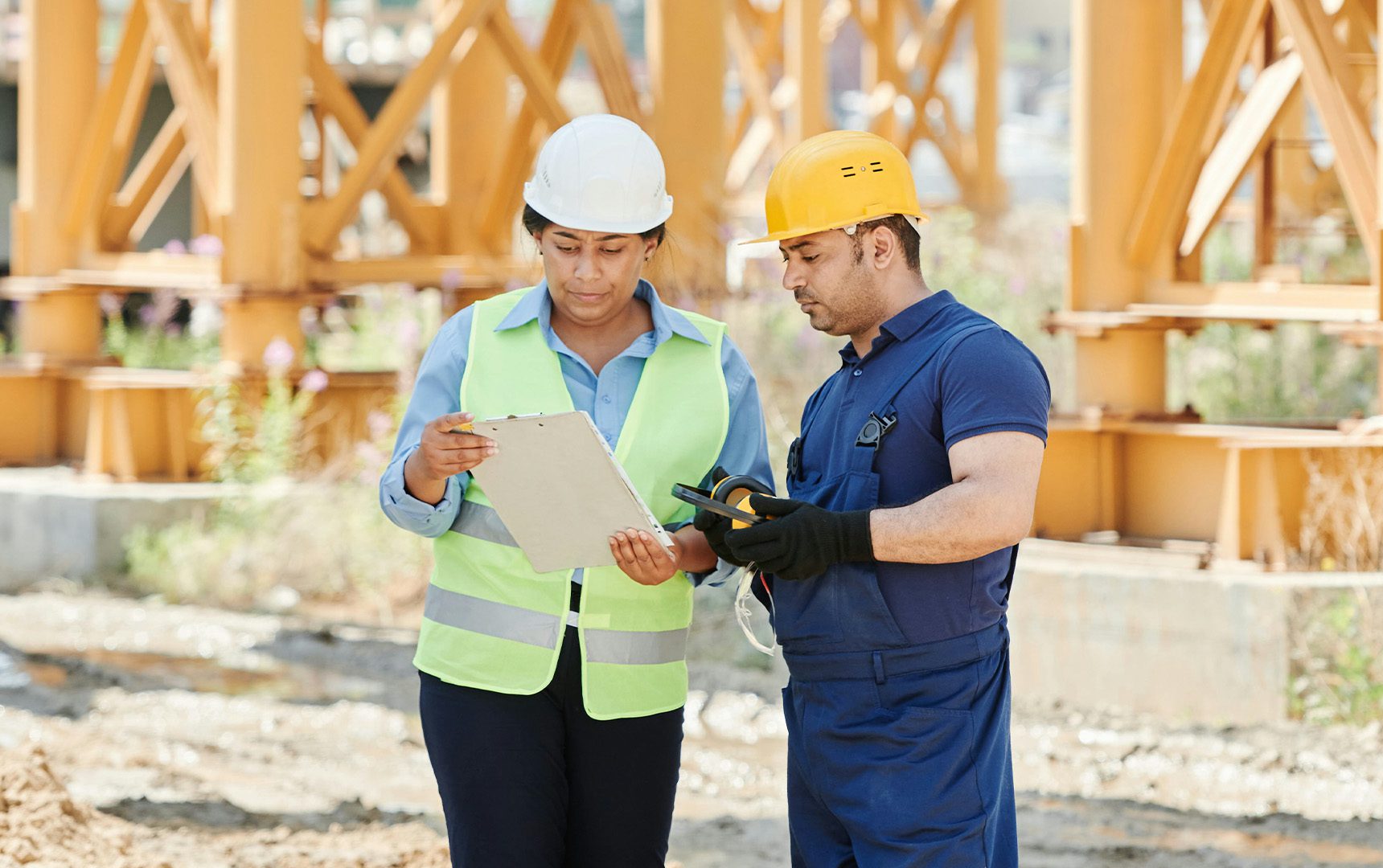 Two construction workers wearing safety gear reviewing a clipboard and measuring tool on a jobsite, with structural steel beams in the background.