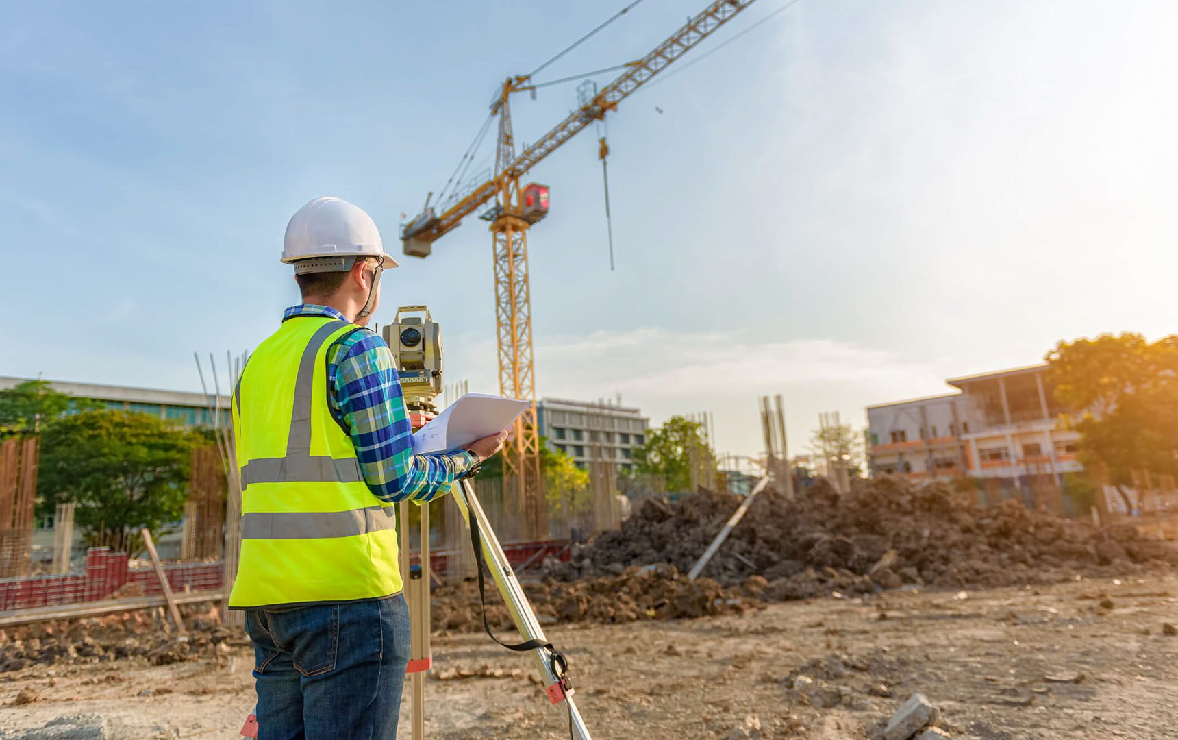 Construction site engineer using surveying equipment and reviewing plans, with a tower crane and building framework visible in the background under a bright sky.