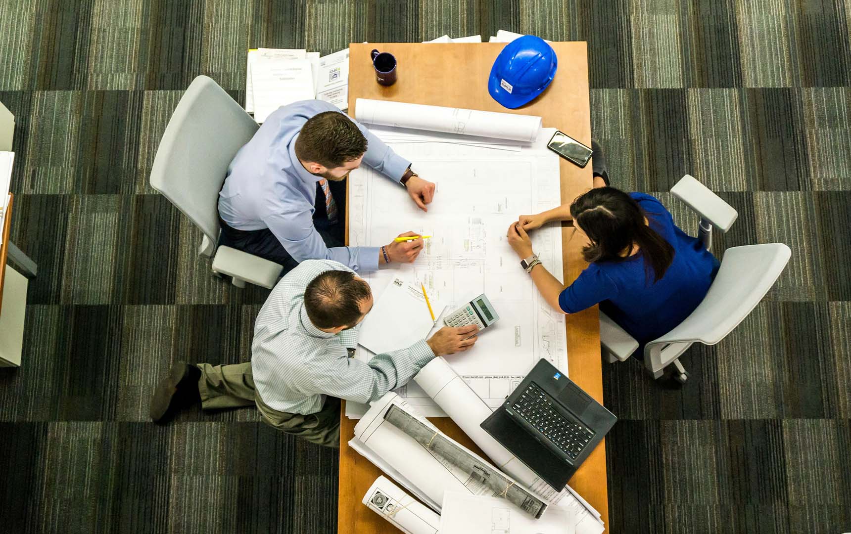 Three professionals gathered around a table reviewing blueprints and using a calculator during a construction planning meeting, with a blue hard hat and laptop nearby.
